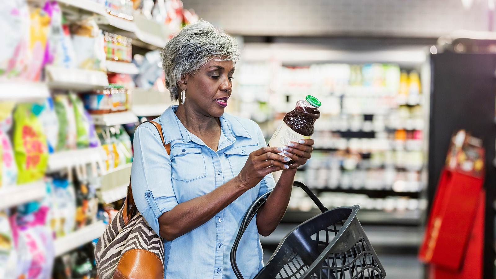 An elderly woman inspects a bottle of juice in a grocery store aisle, with various products displayed on shelves behind her.